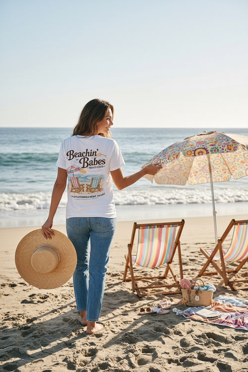 Press Society white cotton t-shirt with 'Beachin' Babes' graphic print and beach scene, worn by a woman on the beach.