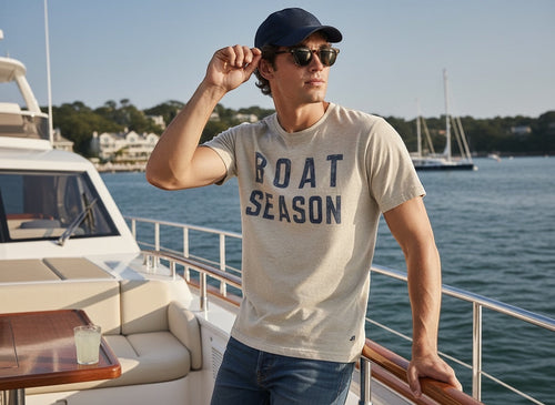 Press Society men's light heather grey cotton t-shirt with navy blue "BOAT SEASON" graphic print, short sleeves, crew neck, worn by a man on a boat.
