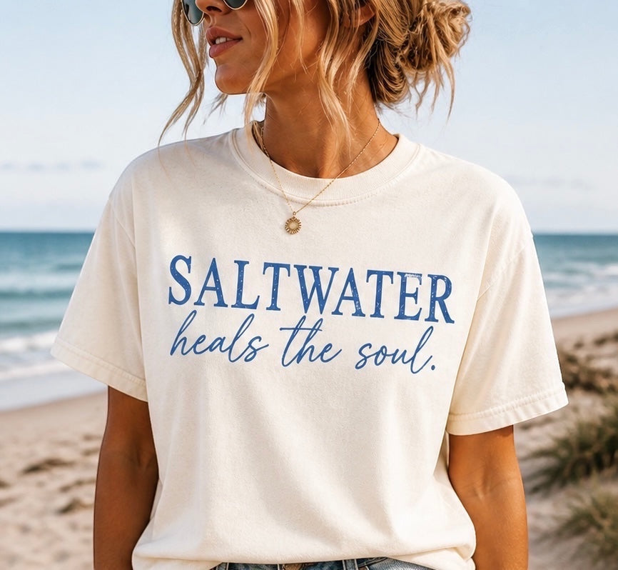 Press Society cream cotton t-shirt with blue "Saltwater Heals the Soul" graphic print, short sleeves, crew neck, worn by a woman on a beach.