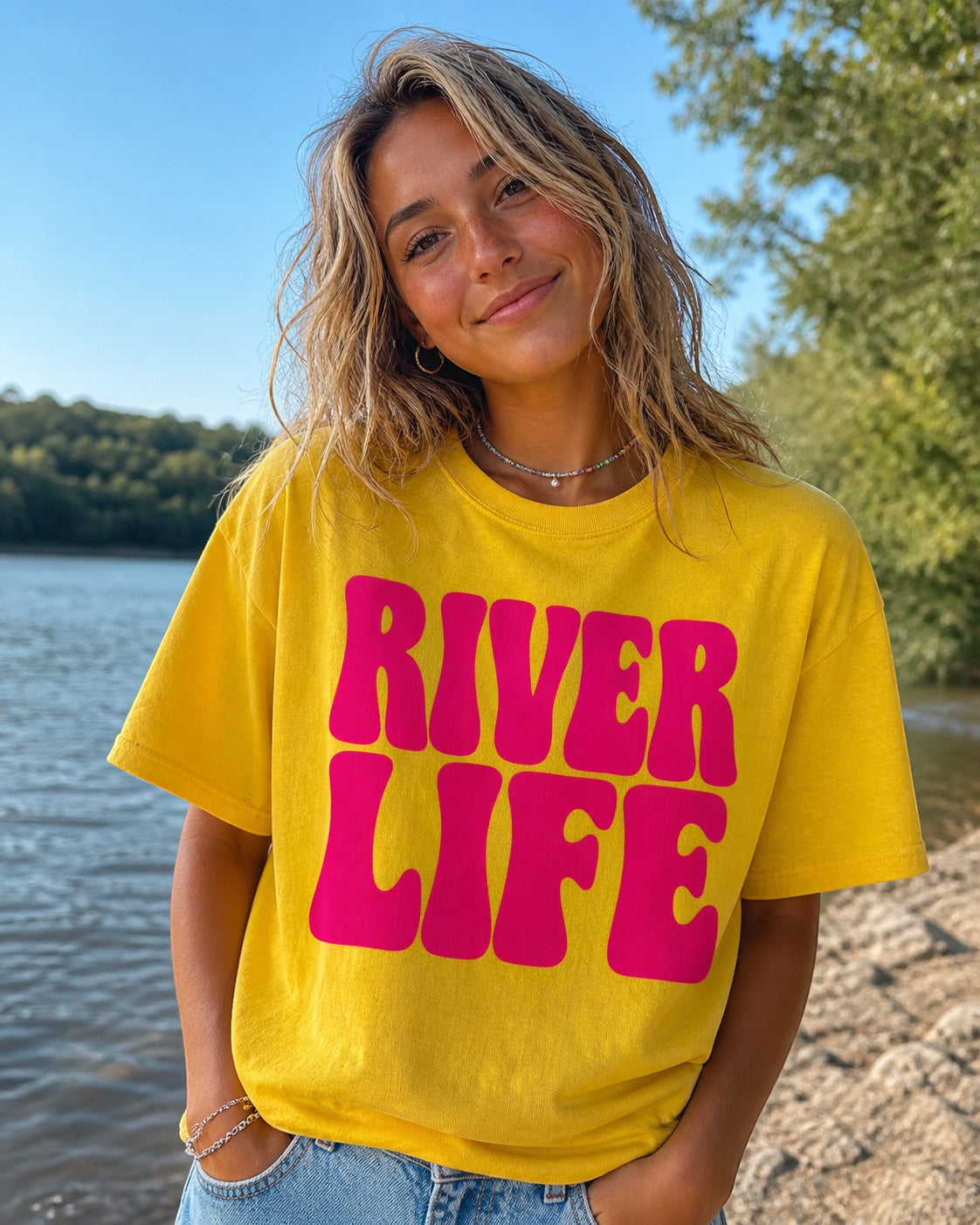 Press Society yellow cotton tee with oversized pink 'River Life' bubble lettering. Model wears it casually by a river.